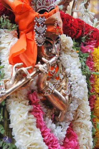 Close photo of a metallic Krishna murti playing a flute, adorned with jeweled crown, pearl and stone necklaces, bangles and an orange cloth draped behind. The idol is surrounded by thick garlands of white jasmine, red and pink roses and yellow marigolds, with green leaves. No living people are visible; the figure is a deity murti, not a person.