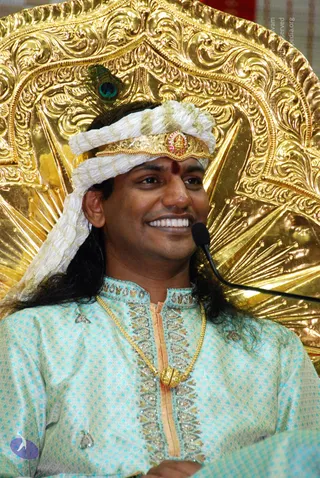 A smiling man with long dark hair sits in front of an ornate gold throne-back with carved floral and sunburst patterns. He wears a white turban with a gold crown-band set with a red stone and a peacock feather, a red bindi on his forehead, a pale turquoise embroidered kurta, and a long gold necklace with a gold pendant. A microphone stands beside him. Swamiji (probable): the long hair, ornate gold crown, turban and ceremonial gold necklace are visible cues, presented here as a seated speaker at a discourse.