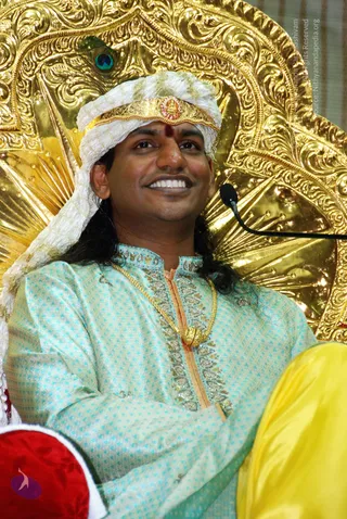 A smiling man seated in front of an elaborate gold repoussé throne backrest decorated with a peacock-feather motif. He wears a white and gold turban with a jewelled central ornament and a red tilak mark on his forehead, long dark hair falling behind his shoulders, a turquoise embroidered kurta with a gold chain necklace, and rests his arms on his lap beside a red cushion and yellow fabric. A black microphone stand is visible at right. Long hair, turban and ornate garments are cues consistent with the candidate named on the page; appearance is only partly distinctive, so identity is uncertain.