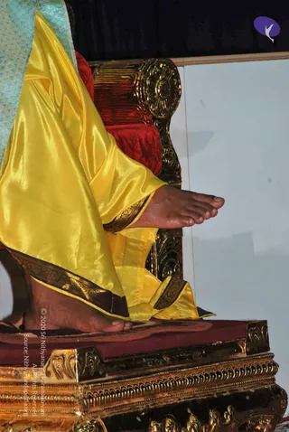 Close-up photograph of a seated person's bare feet and yellow satin garment on an ornate gold-and-red throne. The yellow silk robe has a dark gold-embroidered border, and a turquoise patterned cloth drapes over the knee at upper left. The gilded throne has a red velvet cushion and carved golden scrollwork on the arm and base. Only legs and feet are visible, so no face or identity can be established. A plain white wall is behind the throne.