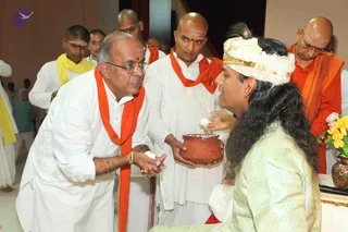 A photograph of a Krishna Janmashtami style gathering indoors. A man in white dhoti and shirt with an orange shawl and glasses leans forward, holding white offering in his cupped hands toward a seated figure. The seated figure wears a white turban with gold trim, a pale green embroidered silk garment, and long dark hair, and is seated behind a low table with yellow and pink flowers in a vase. Behind them, a bald priest in white with an orange shawl holds a terracotta pot containing white material. Several men in white and orange or yellow shawls stand behind, some with tilak marks on their foreheads. A portrait of the seated figure is not shown; the scene is a live ritual exchange.