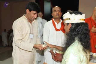 Indoor event photograph of a long-haired man with a white crown band trimmed in gold, wearing a pale green embroidered kurta, offering something from a copper vessel to a man in a cream kurta with a name badge who holds out his cupped hands. Behind them stand a bald man in white with an orange-red shawl and a man in an orange top with a beaded necklace, with other people in white clothing in the background. The setting is a plain hall with a dark red curtain. a person: the long-haired figure in the green kurta and crown band, offering from the vessel, is a probable match for a person on appearance cues of long swept hair and a gold-trimmed crown.