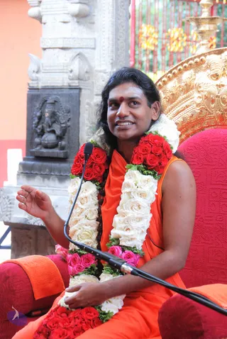A man with long swept-back dark hair and a red bindi sits on a red cushioned throne with ornate gold carved back, speaking into a microphone. He wears an orange sleeveless garment and is draped with long garlands of red and white roses with green leaves, one held in his hand. Behind him stands a grey stone carved shrine pillar with a deity murti in a niche and a red and gold metal gate with brass lamps. He is seated as the speaker at a satsang-style discourse.