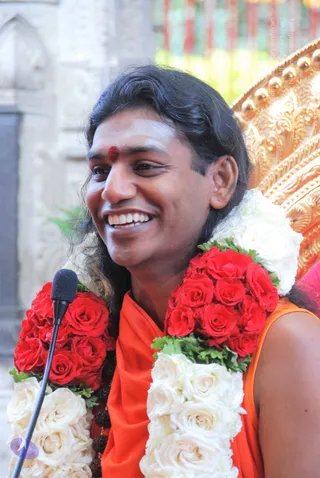 A smiling man with long dark hair swept back sits before a microphone on a stand, wearing an orange-saffron garment and a red bindi on his forehead. He wears thick garlands of red roses and white roses with green leaves draped over his shoulders, and rudraksha beads show near his chest. Behind him is an ornate gold-coloured throne with rounded finials. Swamiji (probable): the long swept-back hair, saffron clothing and rudraksha beads match the page's reference to Swamiji speaking at this satsang.