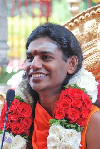 A smiling man with long, swept-back dark hair and a red bindi on his forehead sits in front of an ornate gold throne, wearing an orange saffron garment and a thick garland of red and white roses. A black microphone on a stand is at the left edge. In the background are blurred red, yellow and green decorations. Appearance-based cues including long hair, saffron clothing and the ornate rose garlands are consistent with Swamiji (probable), seated and being honoured.