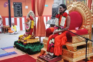 A man in orange robes sits on an ornate gold-and-red throne with a microphone stand in front of him, smiling broadly, wearing long garlands of red and white roses and a rudraksha-style neck. He is bare-footed on a small red mat. Beside him stands a decorated deity murti dressed in a garland of white flowers and yellow and red blossoms, with a green silk cloth draped beneath it on a golden base. A seated man in white robes and a yellow shawl sits to the left near a low table holding a flower-filled tray. Behind them hang an orange curtain and a dark wooden board with a list of names on a striped orange and white wall.