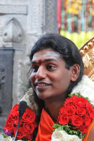 Close-up photo of a smiling man with long, swept-back dark hair and a red kumkum dot with white vibhuti on his forehead. He wears an orange saffron garment and is draped in thick garlands of red roses and white flowers. A black microphone on a stand is in the lower left. Behind him are a weathered stone wall with carved arch detailing and a golden ornate chair edge with colourful hanging decorations in the blurred background. Swamiji (probable): the long hair, saffron clothing and rudraksha-style forehead marking are the cues for this candidate, shown speaking at a satsang setting.