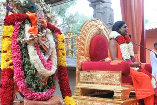 A photograph of a Krishna murti decorated with garlands of red roses, yellow marigolds, white flowers, pink petals and green leaves, with a peacock feather in its crown. Beside it, a man with long dark hair sits on an ornate gold-and-red throne, wearing an orange robe with a red-and-white floral garland, speaking into a microphone. Another person in white stands at the right edge, and a gold railing and pillar are visible behind. The scene appears to be a satsang discourse in an ashram setting. Appearance-based candidate, identity uncertain.