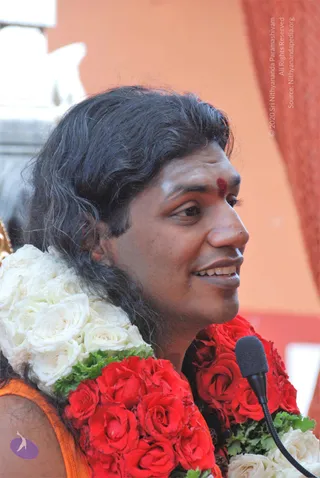 A close-up side-profile photo of a smiling man with long, swept-back dark hair and a red kumkum mark on his forehead, speaking into a black microphone on a stand. He wears long garlands of red roses and white roses across his shoulders, with green leaves between them, and an orange garment is visible at his shoulder. The background is a warm orange wall. a person: the long swept hair, red forehead mark, garlands and orange clothing suggest he is the discourse speaker at this satsang. A white-and-orange wall and a microphone indicate a discourse setting.