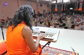 Seen from behind, a person with long dark curly hair in a sleeveless orange top sits on a red cushion at a white electronic keyboard, playing with the right hand. In front of them, a large hall holds a seated audience of many people in white clothing, some wearing white turbans, with white-shirted camera operators and tripods near the stage. Above, multicoloured cloth banners hang from the ceiling, and a small screen is visible at the back. A patterned red-and-cream rug covers the stage floor, with brass items and a peacock feather nearby.