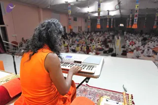 A photograph taken from behind a performer seated on a raised white stage, playing a white electronic keyboard. The performer wears a sleeveless orange robe and has long, dark, wavy hair falling past the shoulders. In front of the stage, a large hall is filled with seated devotees in white clothing, some in orange and yellow. Colourful cloth banners in red, yellow and blue hang from the ceiling. A red and gold cushion, a small brass lamp and a flute sit beside the keyboard on a patterned red carpet. A man in white with a yellow shawl stands at the front of the hall, facing the audience.