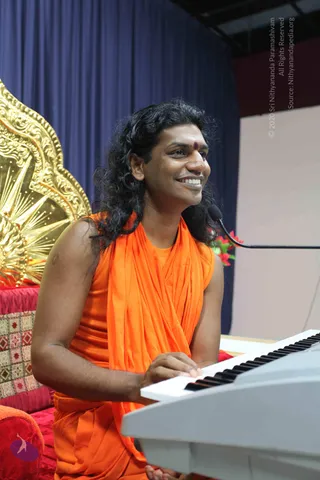A smiling man with long, swept-back dark curly hair and a small red bindi sits behind a white electronic keyboard, playing with his right hand. He wears a sleeveless saffron-orange wrap over his shoulders. A microphone on a stand is at right. Behind him are a navy blue draped curtain and a large ornate gold throne-back with red cushions at left.
