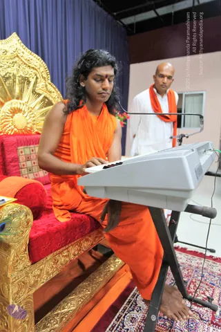 A man with long, swept-back dark hair and a red bindi sits on an ornate gold-and-red throne, playing a white electronic keyboard on a stand. He wears a sleeveless saffron-orange robe and is barefoot on a patterned rug. Behind him stands a bald man in white clothes with an orange shawl, looking toward the keyboard player. A microphone on a boom arm is positioned near the keyboard. A blue curtain and a gold-patterned throne back fill the background.