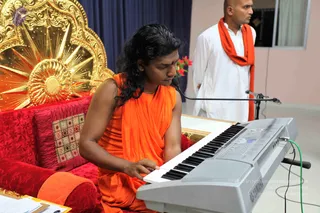A seated figure in saffron-orange robes with long, swept-back dark hair and a red tilak on the forehead plays an electronic keyboard with both hands, sitting on a red velvet cushioned seat in front of an ornate gold sunburst backrest. A microphone on a stand is in front of the keyboard. Behind, a second man in white kurta and a saffron-orange shawl stands near the wall. Swamiji (probable): the seated musician shows long swept-back hair, saffron-orange clothing and a red forehead mark, and is seated on a throne-like seat, matching the page's note that Swamiji was at this event.