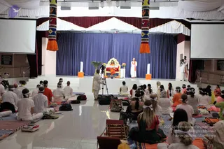 A wide view of a discourse hall with a seated audience on mats facing a stage. Dozens of devotees sit cross-legged, many wearing white clothes and white forehead bands, while some wear orange and red. On the stage, a figure in orange robes sits on an ornate gold and red throne in front of a blue draped backdrop, with a small table holding flowers and a bowl. A second man in white robes with an orange shawl stands at the side, and a cameraman films with a tripod-mounted camera near the front. Decorated pillars with orange tassels flank the stage, and screens hang on both sides.