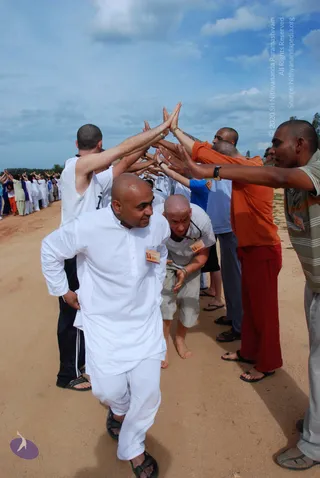 Outdoor photo on a dusty dirt path under a partly cloudy blue sky. A shaved-headed man in white kurta and pyjamas walks forward through a corridor formed by many people raising their arms and palms together overhead. Behind him a man in an orange top and maroon-red trousers and a man in a grey-striped shirt raise their hands, and a bald man in a white t-shirt and khaki shorts with a beaded necklace bends forward. Participants in white and blue clothes line the path in the background. Several wear name badges on lanyards.