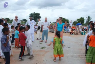 An outdoor photo of a sandy open ground where children play a circle game among adults. A young girl in a green and orange dress stands at the center, with several boys in shirts and shorts around her. A bald man in a white shirt and light blue trousers and a woman in a turquoise top and orange skirt smile nearby. A man in white kurta with a long beaded necklace stands at left-center. Behind them, a seated audience in mixed casual clothing watches under a cloudy sky, with palm trees and a brick wall in the background.