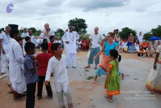 Outdoor photo of a playful group activity on a dusty clearing under a cloudy sky, with a row of seated spectators and trees behind. A woman in a turquoise top and teal trousers with an orange wrap dances barefoot beside a girl in a green dress with orange trim. Several boys in white kurtas and checked shirts gather around them, and a bald man in white clothes with a rudraksha-style mala stands nearby. Other people in white and pastel clothing sit on the ground under a blue umbrella.