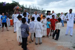 Outdoor photo on a dusty reddish ground in front of an unfinished concrete building with exposed rebar. A colourful striped canopy on a pole appears at the left edge. Several boys in white kurtas, white dhotis and sandals stand in a loose circle with a small group of adults. A boy in a red shirt and a boy in a teal shirt are visible near the center. A man at the center-left in a white kurta with a red-and-gold bead mala stands among the children, and a man on the right in white kurta and white dhoti wears a beaded mala and a name badge. A man in a blue t-shirt and dark shorts stands at the far left. A figure in a flowing white garment with a white shawl is in the middle of the group.