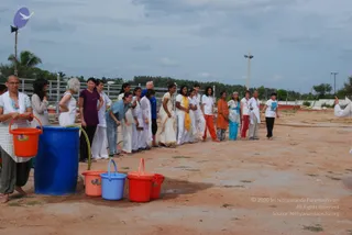 A photograph of a line of women standing on bare dirt ground outdoors, passing buckets in a bucket-brigade style. In the foreground a woman with a shaved head and white shawl over her shoulders holds an orange bucket beside a large blue plastic drum. Other women in white, cream and orange saris and salwar kameez, plus a woman in a purple top and a man in a blue shirt, stand in a row, with red, pink and blue plastic buckets placed on the ground. A white-clad figure appears at the far right edge. A metal fence, palm trees and overcast sky form the background, and a water hose runs into the blue drum.