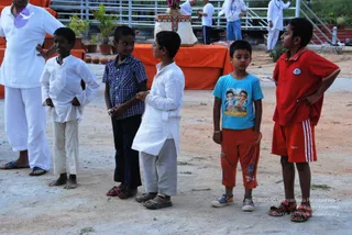 Outdoor scene on a sandy ground with five boys standing in a line. On the left, a man in white shirt and white trousers with flip-flops stands partly visible. Among the boys, one wears a white kurta and light trousers, one wears a blue checked short-sleeved shirt with navy trousers, one wears a light blue t-shirt printed with Bal Ganesh and orange shorts, and one wears a red polo with red shorts. Behind them, an orange-draped altar with a decorated object and potted plants, and people in white clothes stand in the background.