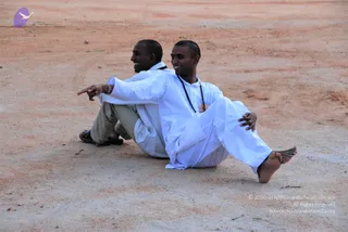 Two smiling men in white kurta-style clothes sit back to back on a dusty reddish-brown ground, one extending his arm and pointing into the distance. The man in front wears a dark rudraksha-style mala around his neck and a small badge on his chest, and is barefoot with his legs stretched out. The man behind wears grey trousers and black sandals. A small purple bird logo appears in the upper left corner.
