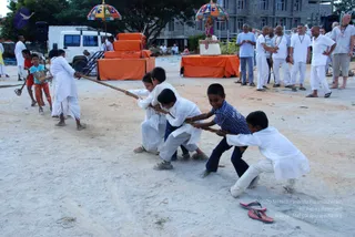 Outdoor photo of children playing a tug-of-war on a sandy ground. Four boys in white kurta-style clothes and one boy in a blue checked shirt pull a thick rope, leaning back with feet planted. Other children in white and a boy in red shorts stand near the rope. Behind them, a row of adults in white kurtas and light clothing watch from the side. Beyond, an orange-draped platform holds a decorated structure under a colourful striped umbrella, and a white van and a concrete building stand in the background. A pair of red slippers lies on the ground in the foreground.