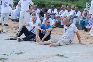 Outdoor photo of a group of devotees seated on a sandy ground and on a low concrete step in front of a red brick wall. Most wear white kurta-style clothes and some have rudraksha malas; a woman on the right wears a light blue head covering and blue dress. Three young men in the foreground sit on the ground: one in a white I Love NY tank top and black track pants, one in a blue t-shirt with a wristwatch, and a shaved-headed man in a white t-shirt with a mala and a badge. A standing man in white clothes stands at the left edge. Several others sit on the steps behind, some smiling and listening. Green grass and a hillside are visible in the background. No visible candidate for a person is confirmed, so the figure is left unsure.