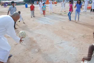 Outdoor photograph on a dusty dirt ground of a playing field. In the foreground a bald man in a white kurta and white dhoti, wearing sandals, leans forward and bends down with his right hand extended, about to kick or roll a white and yellow patterned ball. Behind him a group of children and young people watch and wait: a boy in grey shirt, a boy in red shirt, a small boy in a turquoise shirt and orange-red shorts, a girl in a pink top, and a young woman in a blue top and jeans. Several other people in white clothes stand in the background near parked vehicles and orange containers. A partially visible person appears at the right edge. The scene looks like a casual game of football among ashram residents.
