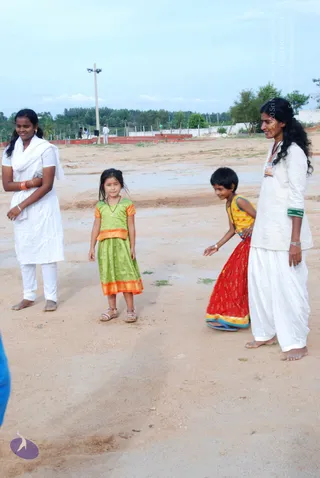 Outdoor photo on a bare dirt ground with a tall light pole, low red-brick walls, trees and a boundary wall behind. Four people stand in a loose group. At left, a young woman in a white kurta and white churidar with a white dupatta stands arms crossed with orange bangles. At centre, a small girl in a green dress with orange trim and sandals stands facing forward. Beside her, a child in a red and yellow skirt with a yellow top bends forward with one hand extended. At right, a woman in a white kurta and white dhoti-style trousers, barefoot, wears a white badge and green bangles. A partial blue figure is at the far left edge.