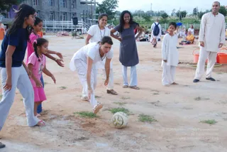 Outdoor photo on a bare dirt ground during a daytime ashram games session. A woman in white top and white trousers bends forward to kick a white-and-yellow soccer ball across the sand. Around her, girls in navy and pink tops and light grey trousers lean in with arms outstretched, and a woman in a grey dress and a woman in white watch and smile. A boy in white kurta stands barefoot at right, and a man in cream kurta and white trousers stands at the far right edge. Behind them, other people in white sit on mats near orange buckets and a white fence, with a multi-storey concrete building in the background.