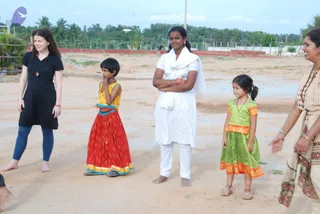 Outdoor photo on a bare sandy ground with a low red brick wall and palm trees behind. Five people stand in a loose line. On the left, a woman with curly hair wears a black knee-length dress and blue leggings, barefoot. Next, a young girl in a red and yellow patterned skirt and yellow top holds her hand to her mouth. A young woman in a white kurta and white trousers, with a white shawl over one shoulder, stands with arms crossed and red bangles. Beside her, a small girl in a green and orange dress and sandals stands with her hair in a ponytail. On the right, a smiling woman in a cream and brown printed sari and bangles reaches toward the group.