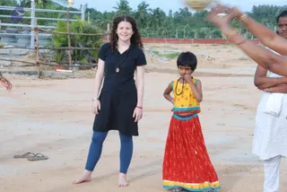 Outdoors on a sandy open ground with palm trees, a red brick wall and a metal fence behind. A smiling woman with curly dark hair in a black short-sleeved knee-length dress, blue leggings and a dark bead mala with a pendant stands barefoot beside a young girl in a red patterned skirt and a yellow-and-blue blouse with a dark bead mala, who stands with her hands together near her mouth. On the right edge, a woman in a white sari-style garment and bangles stands with arms raised, and a bronze-coloured vessel is held up at the top of the frame. Sandals rest on the ground at left.