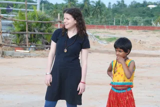 A woman with curly brown hair stands on a dirt ground at a construction-like site with a red brick wall and palm trees behind her. She wears a short-sleeved black knee-length kurta, blue leggings, a dark bead mala with a small pendant, a silver bangle and a red thread on her wrist. Beside her a young girl in a yellow sleeveless choli with orange floral print and a red patterned skirt holds a dark mala near her face and looks off to the side.