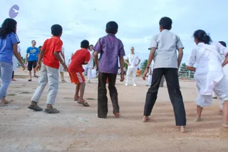 Outdoor photo of children playing a game on a bare sandy ground under a pale cloudy sky with palm trees behind. Several children with their backs to the camera face a central figure: a bald man in a white shirt and white trousers with a white cord necklace, hands on hips, standing at the centre-back. Nearby, adults in white and a blue-shirted man stand around. Children wear a red polo, a purple striped shirt, a grey shirt and black trousers, a blue top, and a white salwar outfit. Sandals and bare feet are visible.