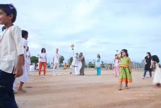 Outdoor photo on a bare dirt ground under a pale cloudy sky during an evening games session. A girl in a green dress with orange trim shades her eyes while looking toward the play area. A child in a cream kurta and navy trousers stands at the left edge. Several adults in white clothes, including an orange-clad figure sitting in the background, watch a ball thrown in the air near a metal fence and palm trees. A woman in a floral sari and another in a blue outfit stand nearby. A few other children in pink and white dresses are scattered around the field, and seated people in white are visible further back.