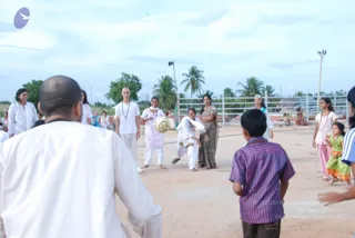 Outdoor photo of a sandy open ground at a community event, with palm trees, a white metal fence and a floodlight pole behind. In the centre, a woman in a white kurta and white trousers, with a white shawl, is mid-motion as she tosses a light ball; several people in white and pastel clothing watch and gather around her. On the right, a young boy in a purple striped shirt and dark shorts faces away from the camera, and a girl in pink and a child in green stand nearby. A bald man in a plain white shirt, seen from behind, occupies the left foreground. Several people in white clothing are scattered in the background.
