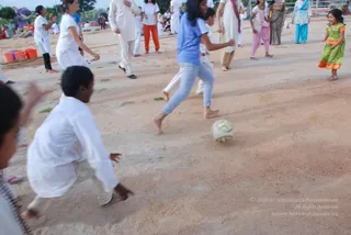 Outdoor photo of a dusty open ground where a game is being played with a white football. In the centre a girl in a blue t-shirt and light blue jeans, barefoot, runs after the ball while a boy in white kurta and white trousers crouches in the foreground, reaching toward it. Behind them several adults in white kurtas and orange and pink salwar kameez watch and move around the field, and a small girl in a green and orange dress laughs on the right. Plastic buckets sit at the left edge and a few people stand near a fence and tent in the background.