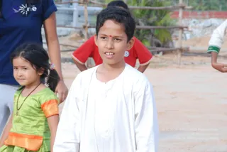 A photograph of outdoors children at a dirt-ground compound. In the foreground a boy with short dark hair and a red bindi wears a plain white kurta and looks to the side. Beside him a young girl with a long dark braid, pink hair tie and beaded necklace wears a bright green dress with orange trim. Behind them a person in a navy blue top stands partly visible, and another child in a red shirt bends forward near a wooden fence and stacked rocks. Only the foreground children are clearly visible.