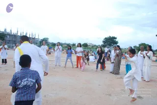 Outdoor photo on a sandy open ground with a partially built concrete structure and trees behind. A man in white clothing with a yellow sash stands at left, facing the group, with his back partly toward the camera. A small boy in a blue checked shirt stands in the foreground. Several people in white and orange clothes watch or move: a woman in a white top and grey-blue trousers stands with hands raised, a girl in pink trousers and a woman in a green sari gesture nearby, and a boy in white leaps at right. A man with a white beard in white clothes stands at far right with hands folded in namaste.