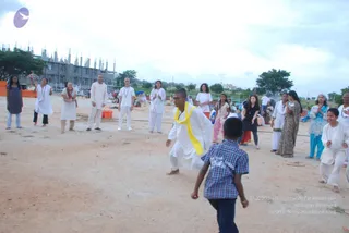 A photograph of an open dusty ground at an ashram-style outdoor event under a pale cloudy sky, with an unfinished concrete building with rebar columns in the background. A man in white kurta and white dhoti with a yellow sash stands in a lunging, mid-motion pose, facing a small boy in a blue checked shirt in the foreground. Around them a loose group of onlookers in white ashram clothing, a woman in a green sari, a woman in a blue salwar, and a woman in pink stand clapping and watching, with several children nearby. Some wear rudraksha-style beaded necklaces and white dress.