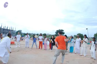 Outdoor photo on a dusty open ground of a group activity. A man in an orange t-shirt and grey trousers in the foreground faces the group, arms bent in motion. Around him, a line of people in white kurtas, white salwar-style outfits, and a few in blue, pink and orange-red clothing stand with hands pressed together or raised. A girl in a blue top and a woman in a light blue salwar kameez are visible. Several wear rudraksha-style beaded necklaces. Unfinished concrete buildings and trees stand in the background under a pale cloudy sky, with a few red buckets and a folded blue mat on the left.