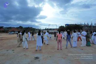 Outdoor photograph of a group of devotees on a bare reddish-brown dirt ground under a cloudy sky, gathered in an open field. Many people wear white kurta or salwar-style clothing, one man wears a yellow sash, a woman in a pink top and pink trousers stands near a woman in a blue top, and a woman in a green sari stands at right. Some figures are in conversation or moving in place. Behind them are low brick buildings, a metal frame structure, parked vehicles, trees and a partly built multi-storey building with rooftop spires. No clear a person candidate is identifiable from appearance.