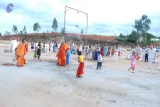 Outdoor photo of a Janmashtami-style pot-breaking game on a dusty open ground. A metal frame holds a hanging clay pot overhead, with a rope dangling from it. Two monks in saffron-orange robes stand near blue barrels, one reaching up toward the suspended pot. Children in bright clothes, including a girl in yellow and red and another in pink, run and play nearby. Behind them a crowd of people in white and colourful garments watches from a brick wall with trees beyond. Water droplets are visible in the air.