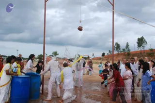 Outdoor Dahi Handi-style celebration on a muddy open ground under a heavy grey sky. A clay pot hangs from a rope strung between two tall red metal poles at upper centre. Participants in white clothes are splashing and pouring water and coloured liquid from blue plastic drums and red and pink containers. A bald man in white is at centre-left, a man in white with a yellow sash stands near the drums, and women in yellow-bordered white saris and a woman in a red top and pink sari are among the crowd. Onlookers stand behind a low orange brick wall with trees beyond.