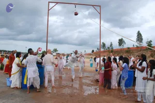 Outdoor photograph of a Krishna Janmashtami celebration on a muddy open ground under overcast sky. A tall rust-coloured metal frame holds a hanging clay pot, with a rope stretched diagonally across the frame. A man in white clothes is mid-leap, reaching toward the pot, while people around him in white kurtas and saris throw water from pink and blue buckets and blue plastic drums. Standing spectators in white, yellow, red and orange clothing watch and cheer along the edges. A blue plastic tub stands at the lower left, and a brick wall and trees form the background.