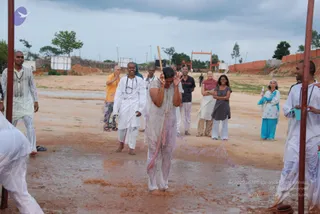 Outdoors on a bare reddish-brown dirt ground after rain, a man in soaked white clothes stands barefoot with arms raised and a wooden stick held upright in one hand, while pink-stained water splashes around him. Several people in white and orange garments stand nearby watching, including a man in orange robes, a man in a white kurta, and women in cream and blue saris and a grey top. Two people on the left and right edges hold metal poles and wear white. Behind them are an orange gate structure, a brick wall, scattered trees and a cloudy sky.