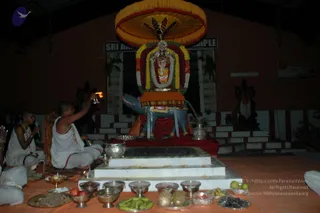 Night-time puja at an open-sided shrine hall with an orange floor cloth. In the centre, a grey-blue Nandi bull stands before a deity murti decorated with marigold and multicoloured flower garlands and an ornate silver-toned face, under a large yellow fringed umbrella canopy. Two shaven-headed priests in white dhotis and gold chains sit on the left; the one on the right raises a lit oil lamp toward the deity while the other chants into a microphone. Silver bowls with bananas, apples, pineapple, coconut and other offerings are arranged on the floor in front. A sign above reads Sri Ar... Temple. Two other seated people are partly visible at the frame edges.