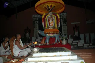 A photograph inside a temple hall at night shows a ceremonial scene centred on a large Ganesha murti seated on a blue-grey elephant-like mount, decorated with a tall yellow umbrella canopy and layered garlands in orange, yellow, green and red. Two men sit cross-legged on a low white platform in front of a raised fire pit with a flame, wearing white dhoti-style clothes, gold-coloured necklaces and shaved heads; the man in front tends the fire while the other sits behind with a hand raised. Silver vessels, a small brass lamp and flower offerings lie on the platform. Behind the murti is a tiered white-and-red step structure with a dark pillared backdrop. The setting suggests a homa or puja.