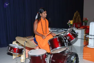 A man in saffron-orange robes with long dark hair and rudraksha beads drums on a red drum kit on a stage. He wears a red-and-white tilak on his forehead and grips two drumsticks, striking the snare and tom. Behind him a keyboard sits on a stand, a gold throne-like chair with a red cushion stands at the right beside flower arrangements, and a dark blue curtain hangs in the background. A white fan stands at the right edge. A second person is partially visible behind the drummer.